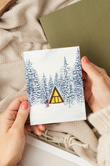 Person holding a card with a winter scene and triangle house illustration on a textured surface.