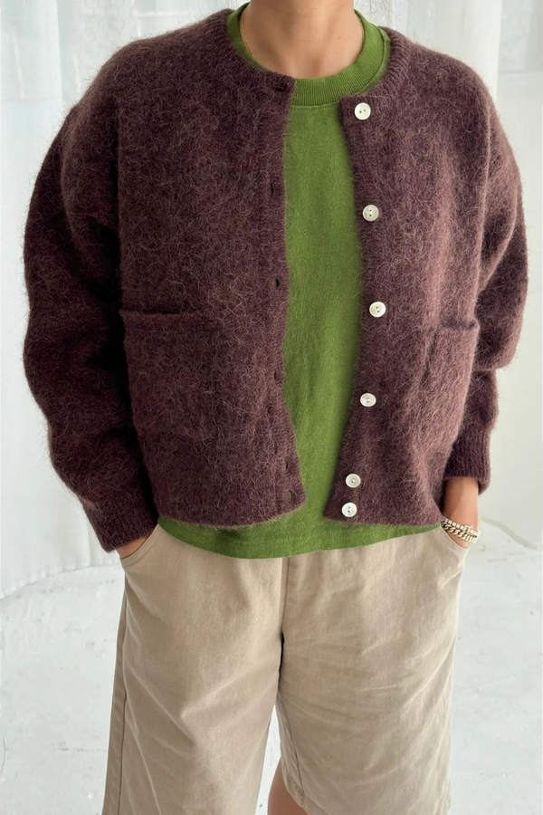 Person wearing a brown cardigan over a green shirt with beige pants on a white background
