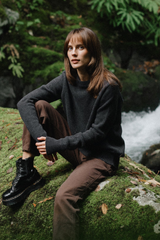 Woman sitting on a rock by a stream wearing a dark sweater and brown pants.