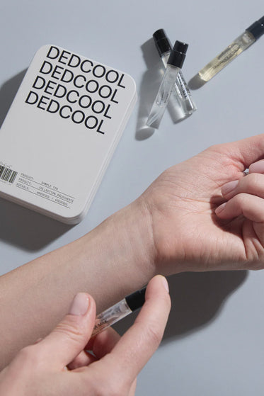 Person applying DEDCOOL perfume product to wrist with a small applicator on a gray background