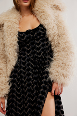 Person wearing a black dress with gold patterns and a beige fluffy coat.