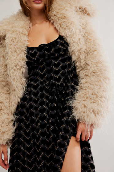 Person wearing a black dress with gold patterns and a beige fluffy coat.