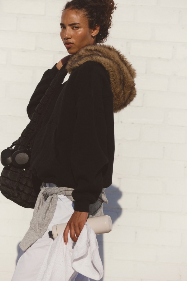 Person wearing a black hoodie with a fur-lined hood against a white wall