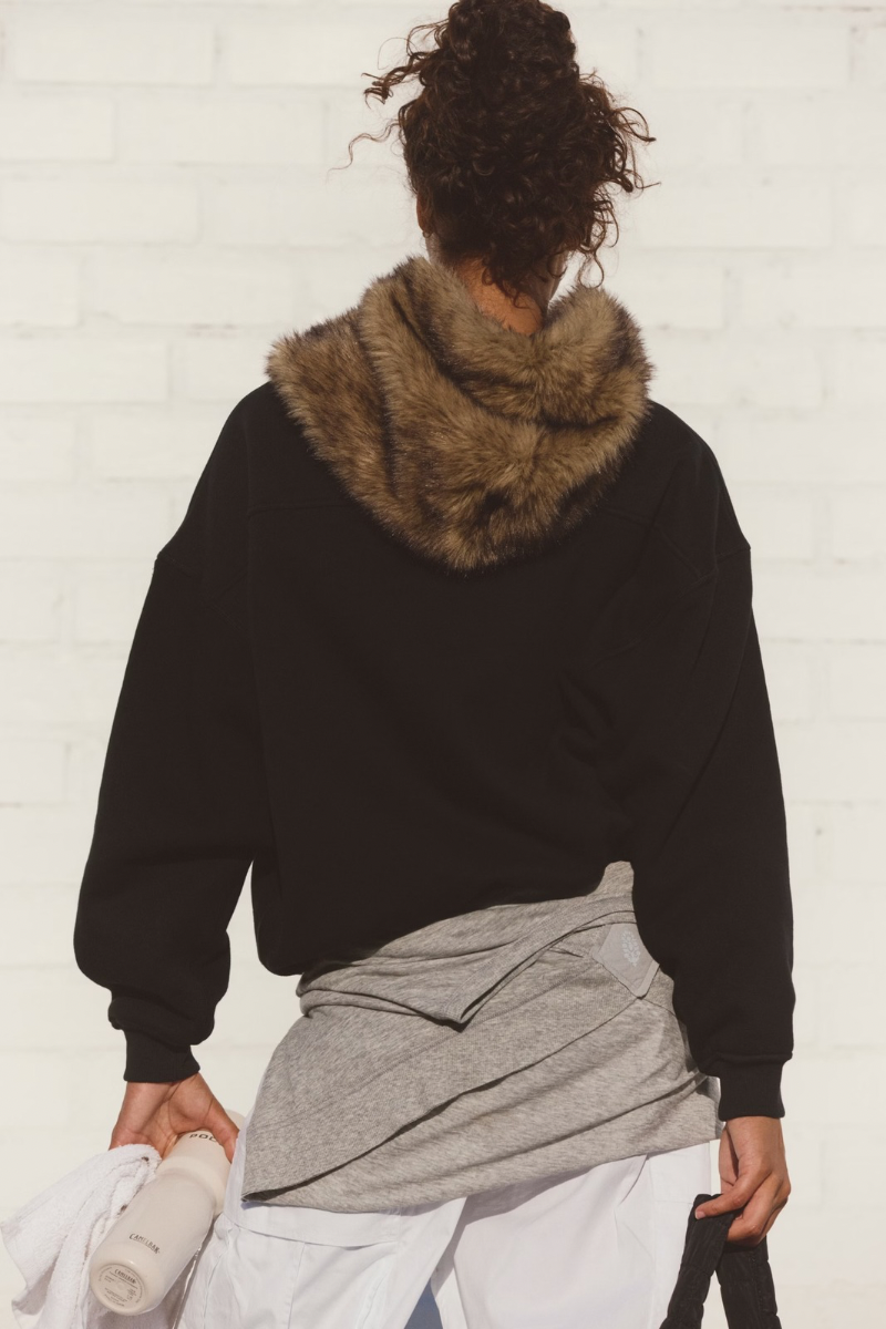 Person wearing a black sweater with a fur-lined hood against a white wall