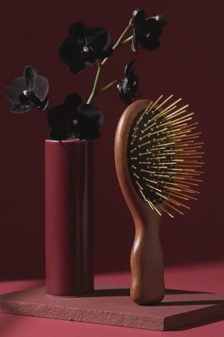 Wooden hairbrush with gold bristles next to a vase with black flowers on a red background