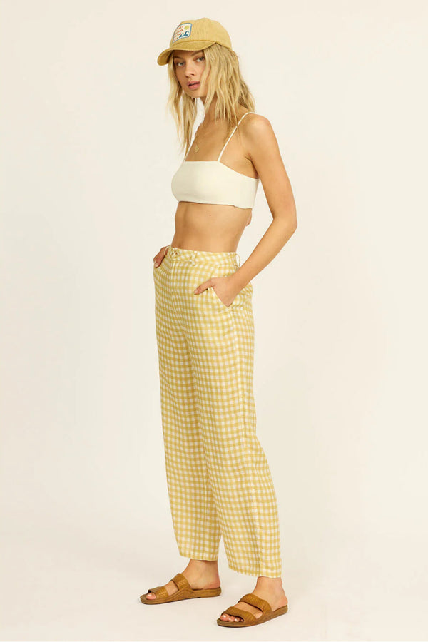 Woman wearing a white top and yellow checkered pants with a beige cap on a light background