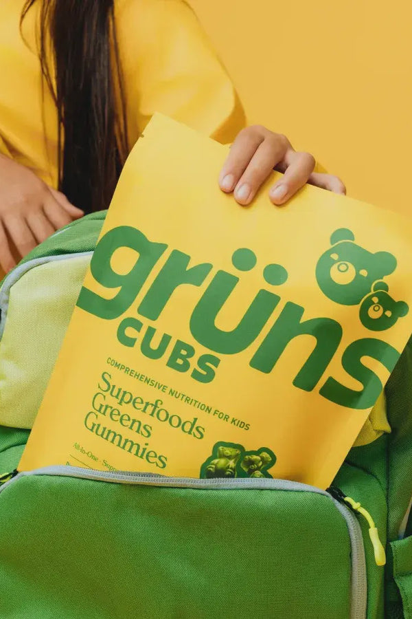 Person holding a yellow 'grüns Cubs' package over a green backpack on a yellow background