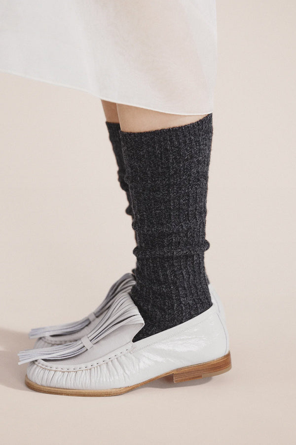 Person wearing dark gray knitted leg warmers with white shoes on a beige background