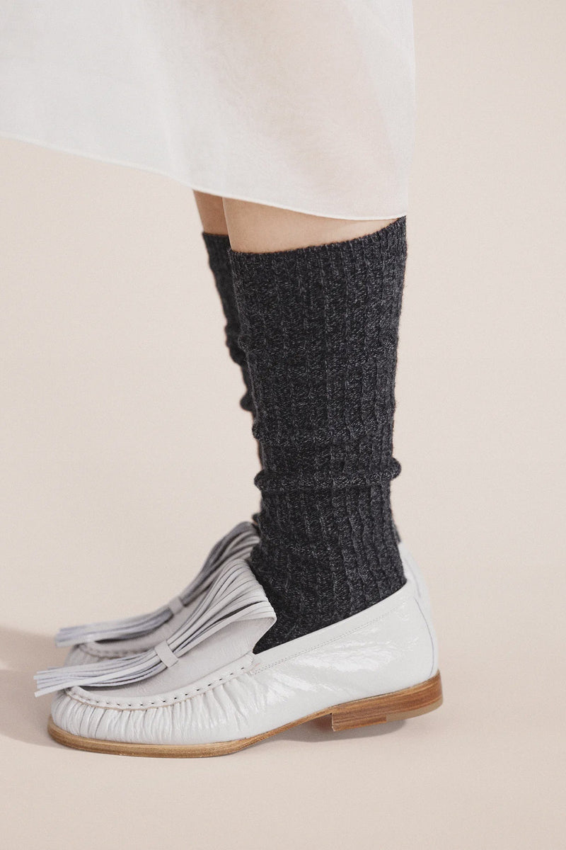 Person wearing dark gray knitted leg warmers with white shoes on a beige background