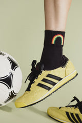 Person wearing yellow sneakers with black stripes, black socks with a rainbow design, and holding a soccer ball on a light green background.