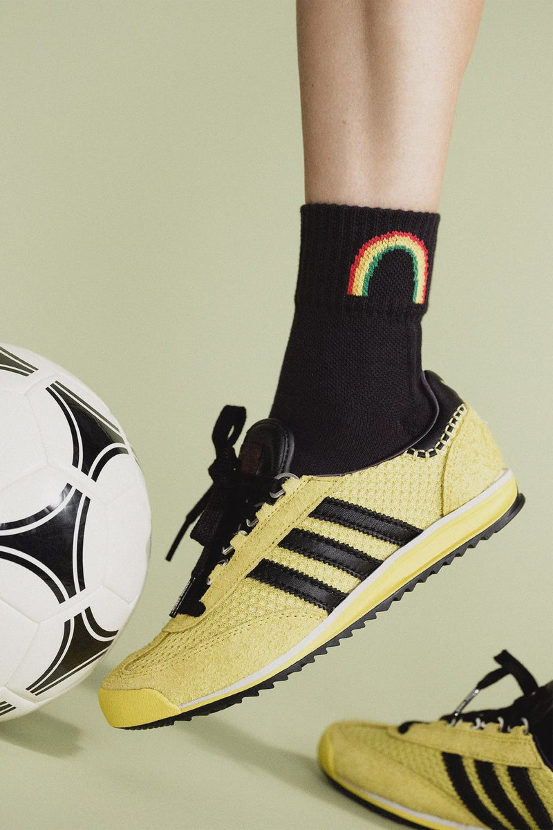 Person wearing yellow sneakers with black stripes, black socks with a rainbow design, and holding a soccer ball on a light green background.