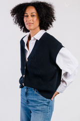 Person wearing a navy sweater vest over a white shirt with blue jeans on a light gray background