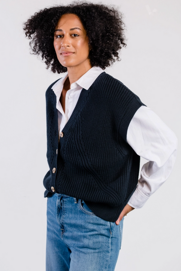 Person wearing a navy sweater vest over a white shirt with blue jeans on a light gray background