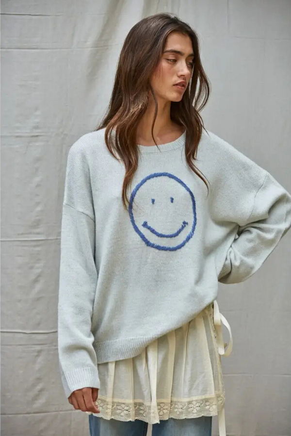Woman wearing a light gray sweater with a smiley face design against a plain background