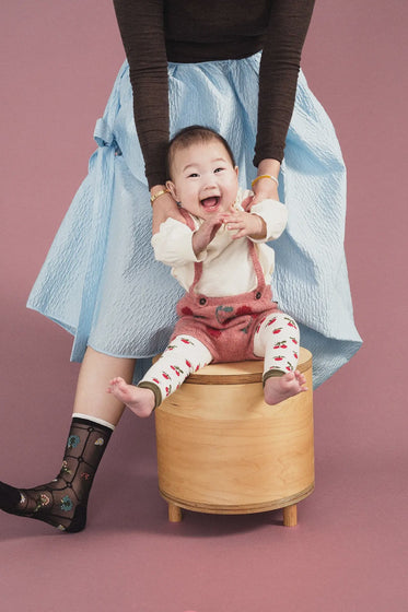 Child sitting on a wooden stool with a person holding their hands, against a pink background