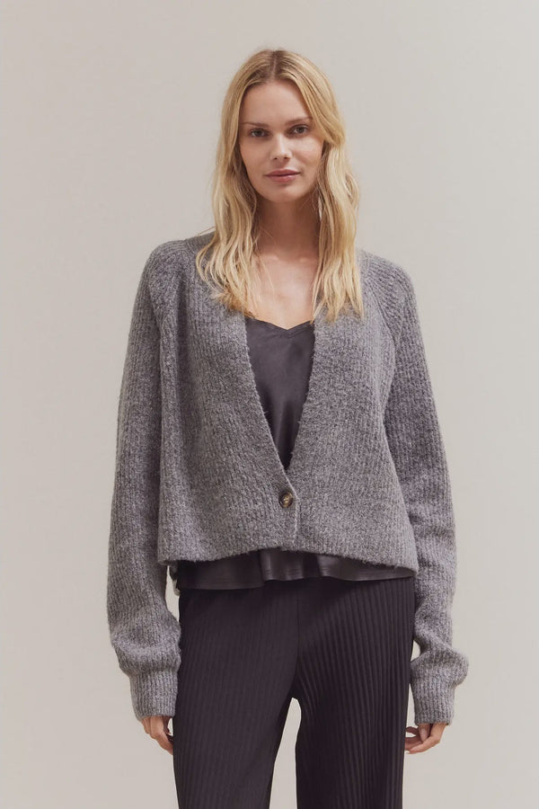 Woman wearing a gray cardigan over a dark top and pants on a plain background