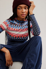Person wearing a patterned sweater and dark pants sitting against a plain background