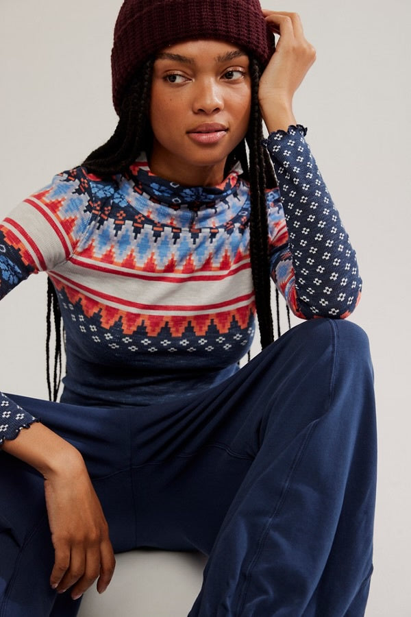 Person wearing a patterned sweater and dark pants sitting against a plain background