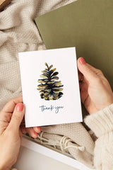 Hand holding a card with a pine cone design and 'Thank you' text, placed on a textured surface.